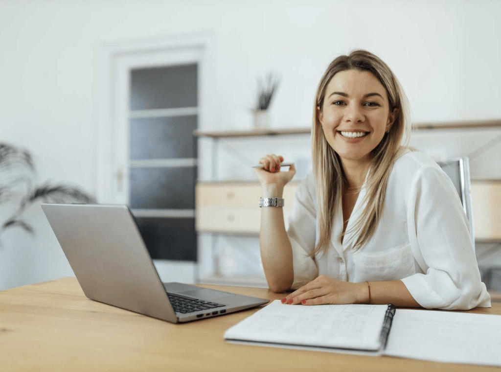 EpicVision Biuro Rachunkowe. O nas. Uśmiechnięta kobieta w białej koszuli siedzi przy biurku z laptopem i notatnikiem, gotowa do pracy.